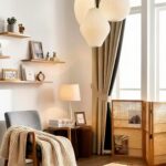 A cozy, minimalist reading nook with a warm, Japandi-style aesthetic. On the left, a grey armchair is draped with a chunky, cream-colored knit blanket. Behind the chair, a small wooden side table holds a lighted table lamp with a glass base, stacked books, and a small framed picture. The white wall features staggered, light-wood floating shelves decorated with small plants, books, a vintage clock, a wooden calendar block, and framed art. To the right, a large window with sheer white curtains is framed by heavy, beige drapes tied back. Hanging from the ceiling are three layered, white paper origami pendant lights. On the floor by the window sits a three-panel woven rattan room divider holding magazines. The entire space is illuminated by soft, warm light.