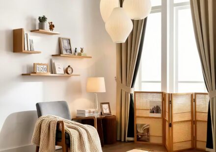 A cozy, minimalist reading nook with a warm, Japandi-style aesthetic. On the left, a grey armchair is draped with a chunky, cream-colored knit blanket. Behind the chair, a small wooden side table holds a lighted table lamp with a glass base, stacked books, and a small framed picture. The white wall features staggered, light-wood floating shelves decorated with small plants, books, a vintage clock, a wooden calendar block, and framed art. To the right, a large window with sheer white curtains is framed by heavy, beige drapes tied back. Hanging from the ceiling are three layered, white paper origami pendant lights. On the floor by the window sits a three-panel woven rattan room divider holding magazines. The entire space is illuminated by soft, warm light.