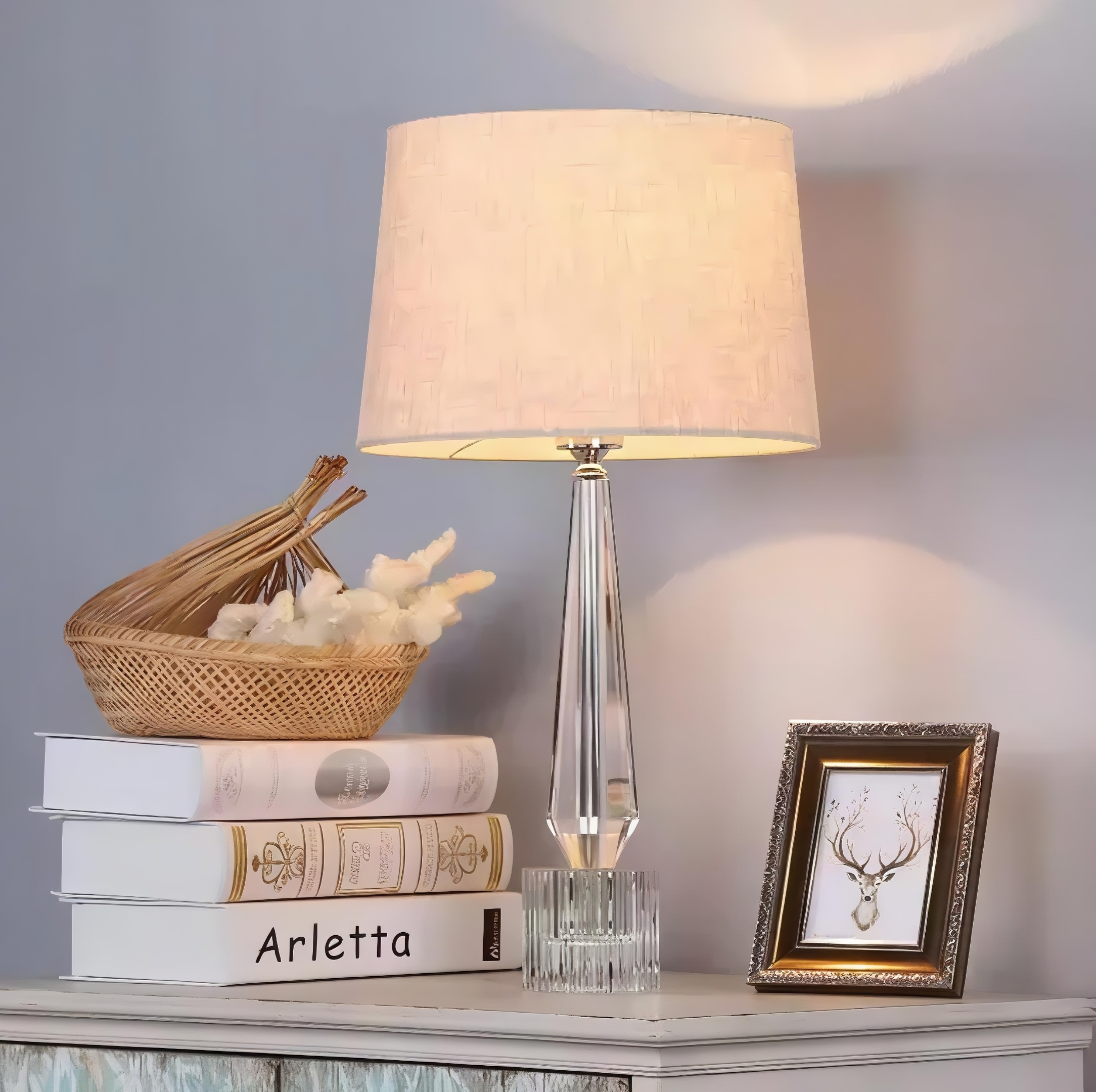 A lit traditional table lamp with a clear, faceted crystal base and a textured cream shade, set on a cabinet next to a stack of decorative books and a framed deer illustration.