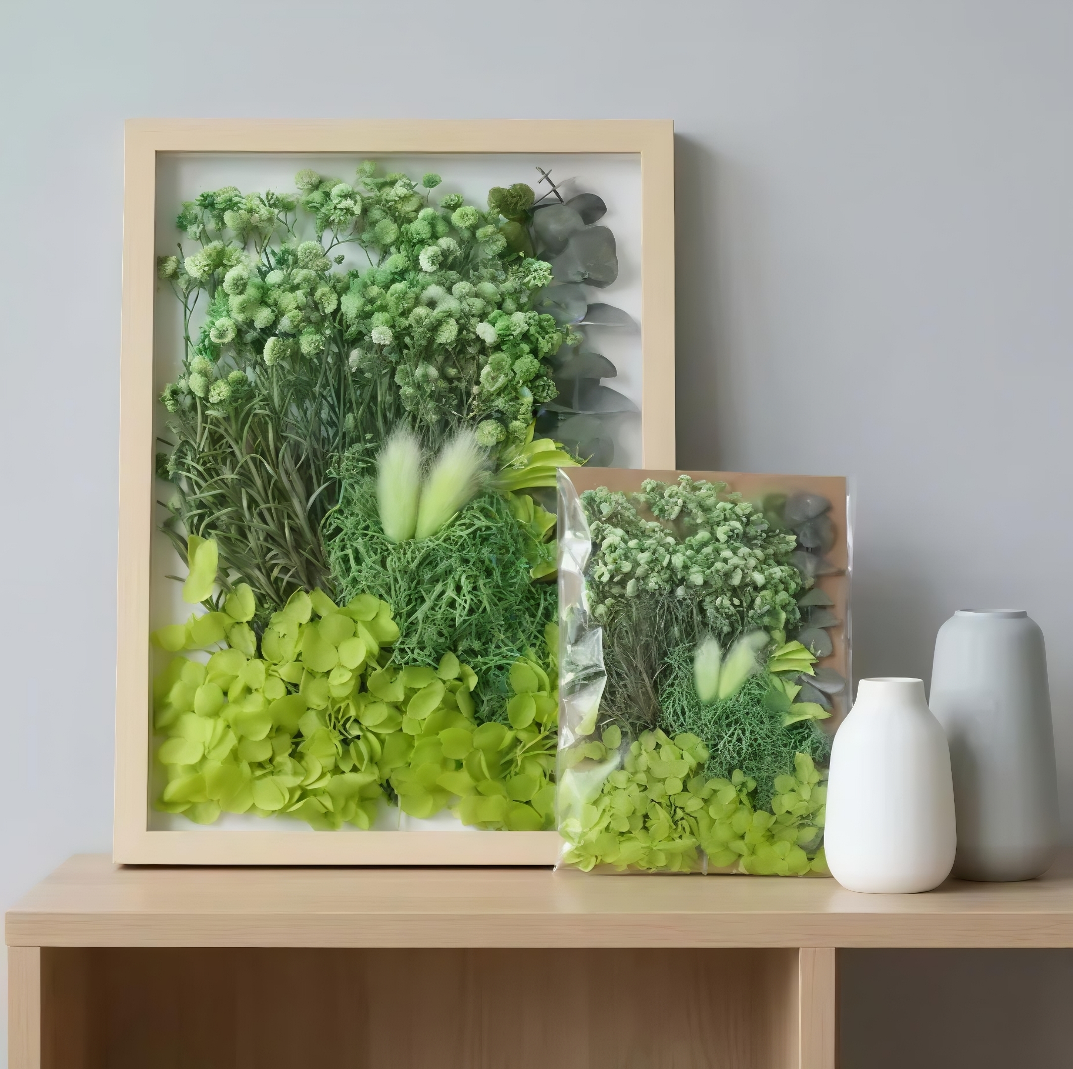 A large and small light-wood picture frame filled with structured arrangements of preserved green moss, ferns, and dried foliage on a wooden surface.