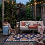 A cozy backyard patio at dusk featuring a wooden pallet sofa with cream and terracotta cushions, a patterned blue area rug, hanging string lights, and a warm brick fire pit zone.