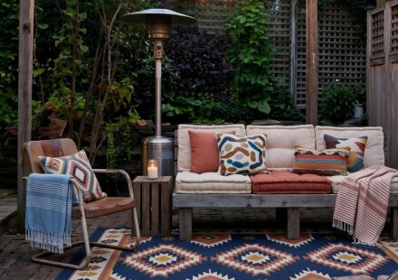 A cozy backyard patio at dusk featuring a wooden pallet sofa with cream and terracotta cushions, a patterned blue area rug, hanging string lights, and a warm brick fire pit zone.