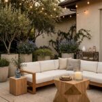 A warm, inviting patio scene at dusk featuring a low-profile cream-colored sectional sofa with light wood trim. In front of the sofa sits a unique, geometric-carved light wood coffee table holding a stack of ceramic plates and a large pillar candle in a glass hurricane vase. The space is illuminated by warm string lights draped overhead. In the background, large neutral stone planters filled with olive trees and purple flowering bushes line a textured stucco wall.